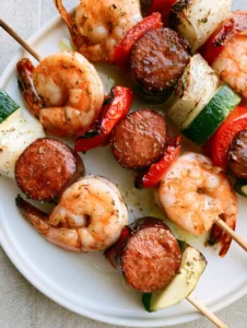 High-angle view of Grilled Seafood Skewers on a white ceramic plate, showing charred edges on smoked sausage and glistening herb oil marinade.