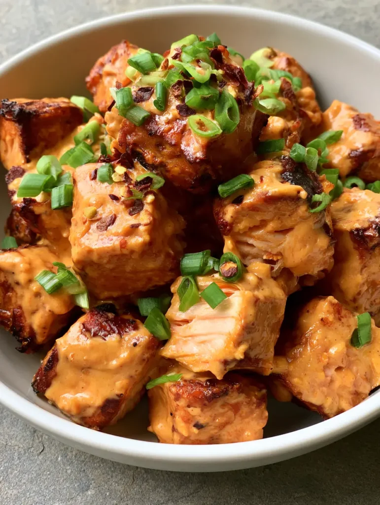 Close-up handheld smartphone photo of bite-sized Bang Bang Salmon Bites with glossy sauce and green onions.