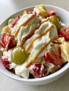 A handheld smartphone photo of a white bowl filled with creamy cheesecake fruit salad and a thin zigzag of amber caramel.