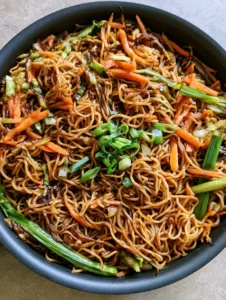 Close up of curly wheat noodles with seared edges and a dark savory sauce glaze in a black skillet. (Homemade Chow Mein Recipe)