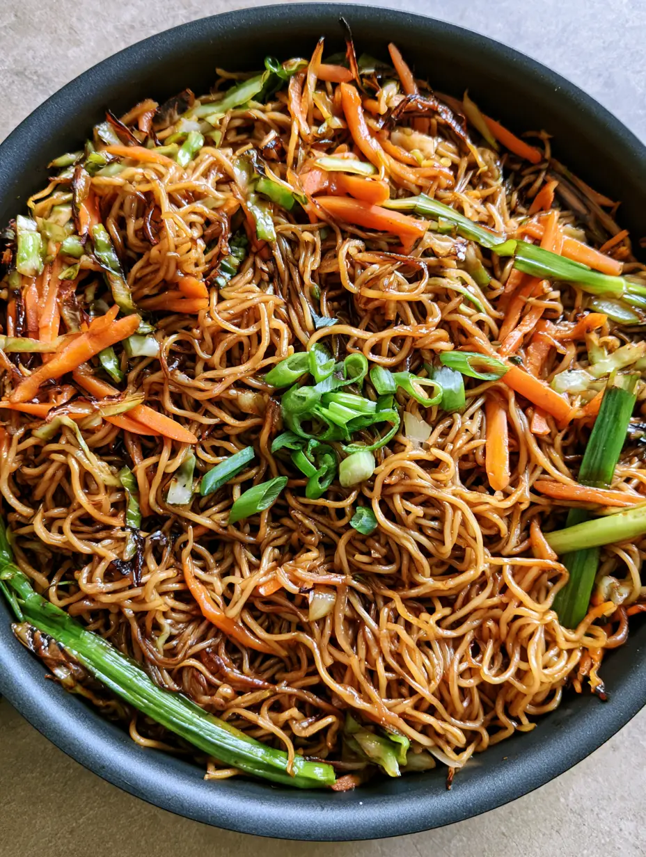 Close up of curly wheat noodles with seared edges and a dark savory sauce glaze in a black skillet. (Homemade Chow Mein Recipe)