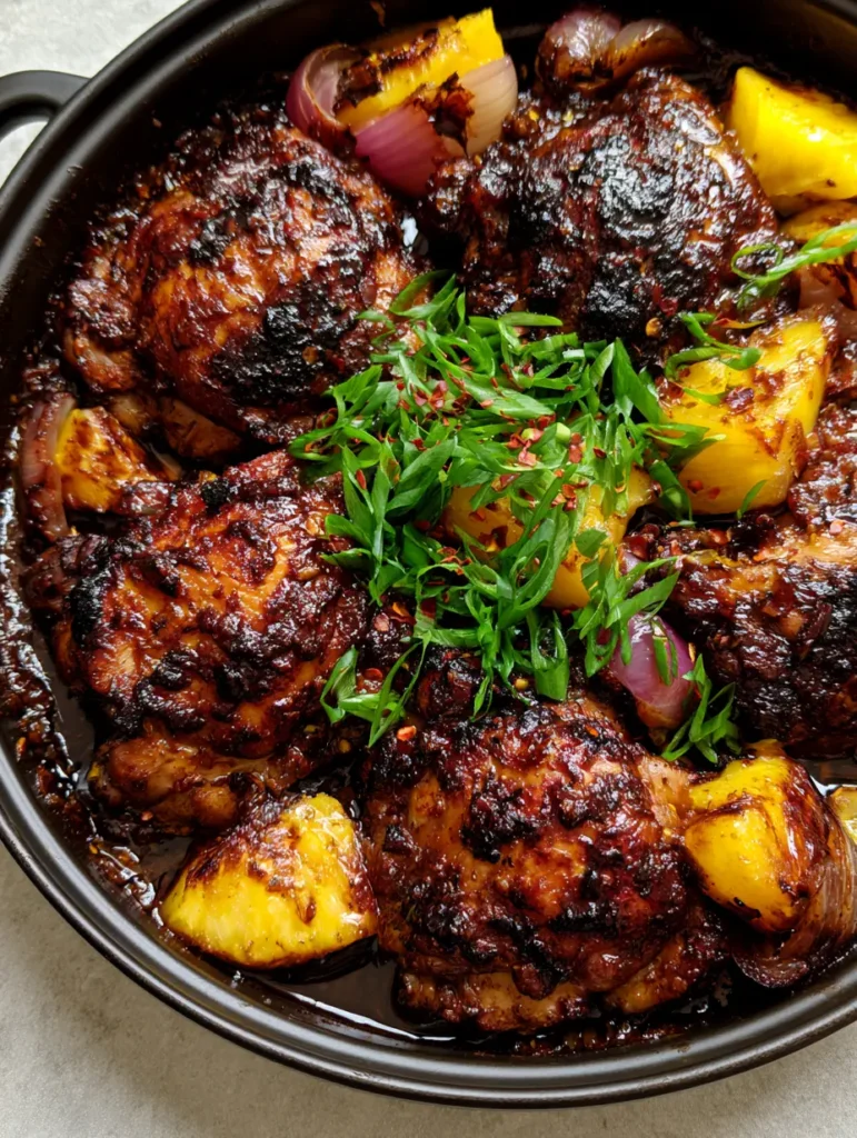 A rustic black baking dish filled with Jamaican jerk chicken, vibrant yellow pineapples, and fresh green scallions.