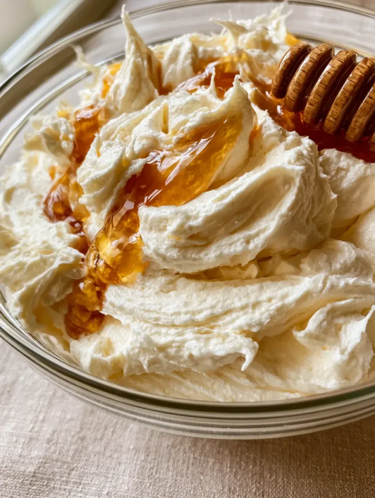 Honey Buttercream Frosting being drizzled with golden amber honey using a wooden honey dipper.